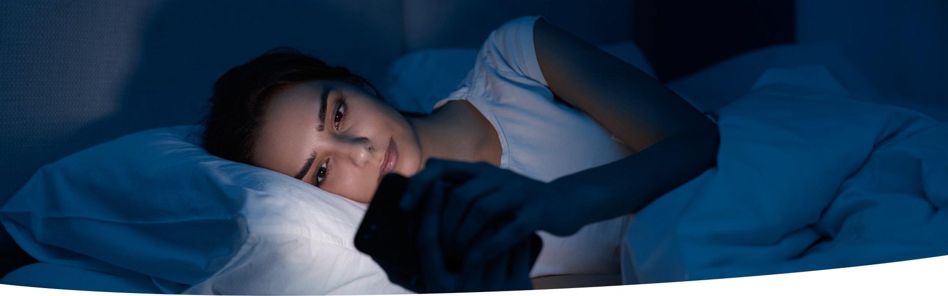 Junge Frau mit Einschlafproblemen liegt mit Smartphone in der Hand im Bett