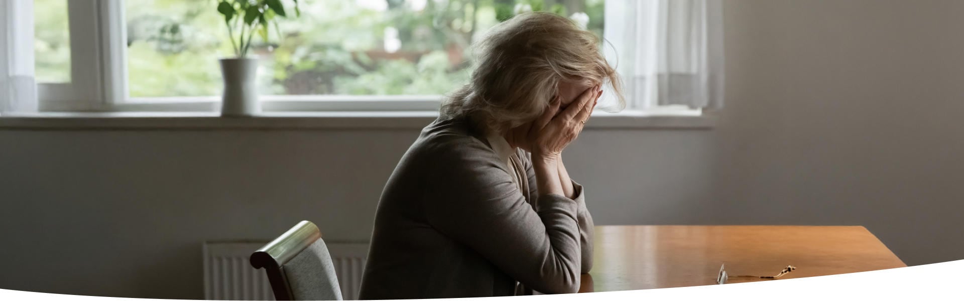 Ältere Frau sitzt am Tisch und verbirgt verzweifelt das Gesicht in den Händen
