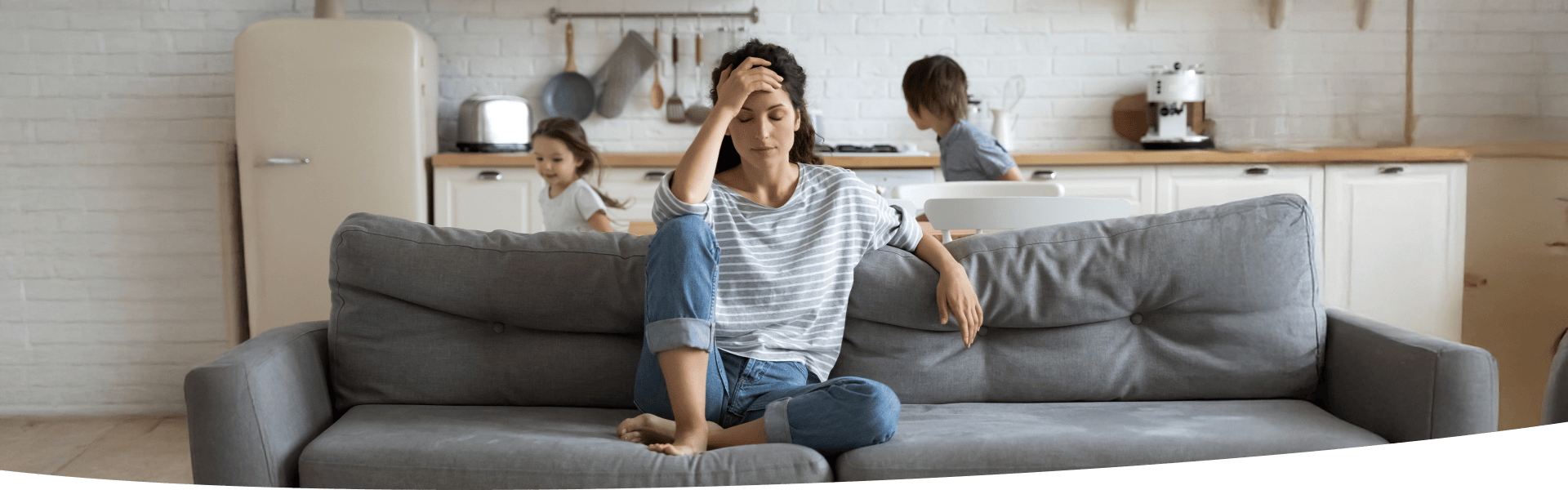 Frau sitzt auf Sofa und hält sich den Kopf, während zwei Kinder um sie herum spielen